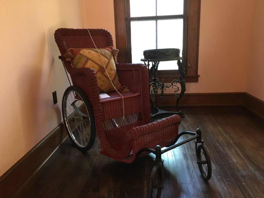 Old fashioned wheelchair. It looks like it's made out of wicker. There is a patterned decorative pillow on it. Next to it, there's a patina looking stand. Behind that, there is an old fashion