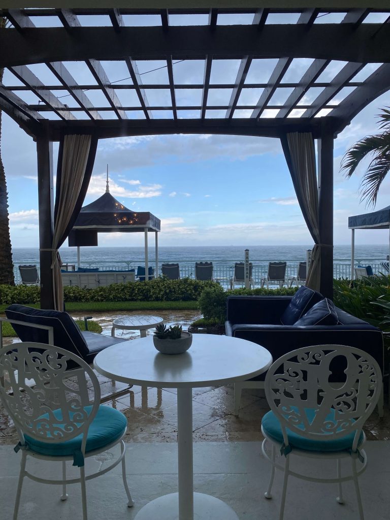 View of the ocean in the background, pool chairs, up close patio chairs, a patio table for two. 