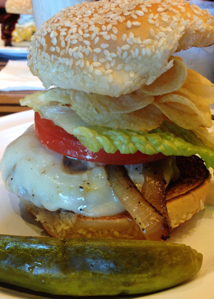 Photo of Nathasha's Burger made at Bobby Flay's Bobby's Burger Palace, burger on a bun, tooped high with cheese, tomato, chips.