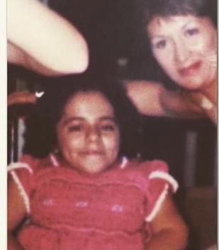 A little girl in a wheelchair who doesn't look 14 but she is. She's wearing a pink plaited dress. Her mom is leaning on the wheelchair handle and they are both looking at the camera.
