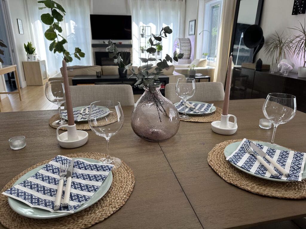 Table setting for four people. Basketweaving placemats. Two long tapered candles. In the middle, a glass jar holding greenery. In the background, the living room. 