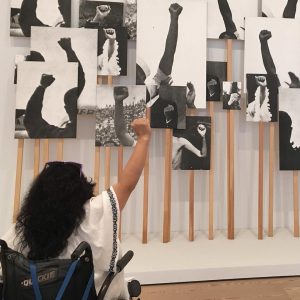 Nathasha's sitting in her black wheelchair. Her back is turned to the camera. Her right arm is held up as a symbol of triumph. She sitting in front of black and white photos with arms extended in triumph. 
