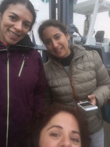 Three latina brunettes. The one on the left is wearing a dark wine parka. The one on the right is a big shorter. She's wearing a dark tan parka. Nathasha is sitting in her wheelchair which isn't in the photo. Only her face from the nose up is in the photo. All three women are smiling at the selfie. 