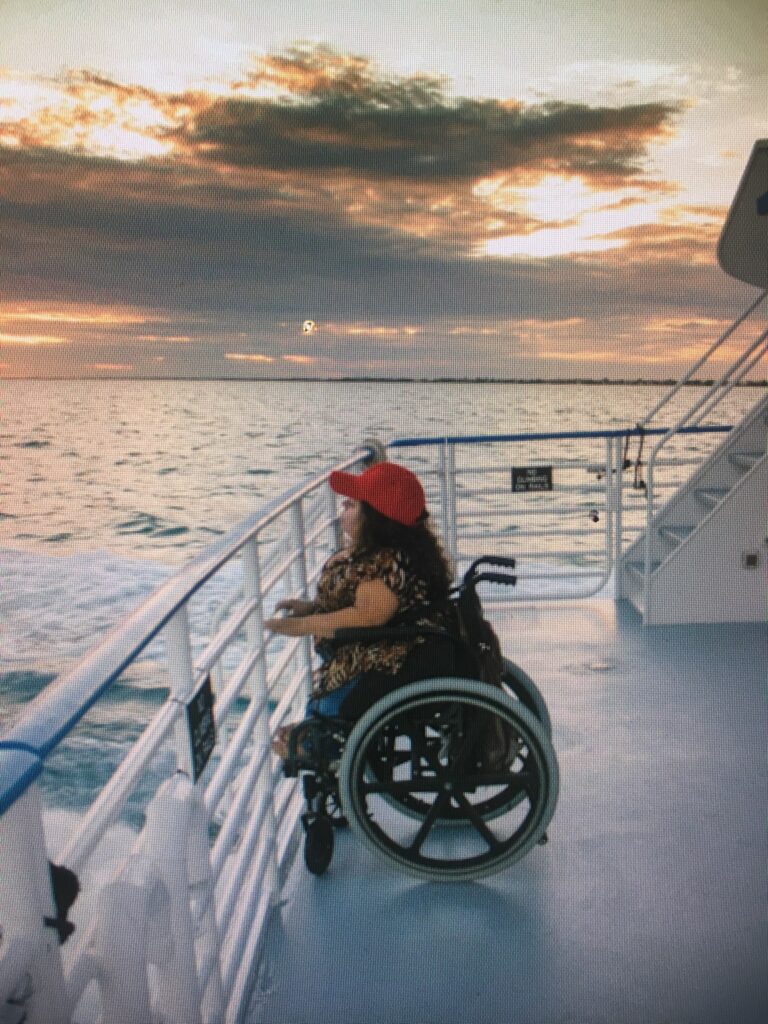 Side view. Nathasha Alvarez sitting in her black manual wheelchair. She's wearing a red baseball cap. Long curly brown hair. She's wearing jeans and animal print short sleeve blouse.She's on a boat
