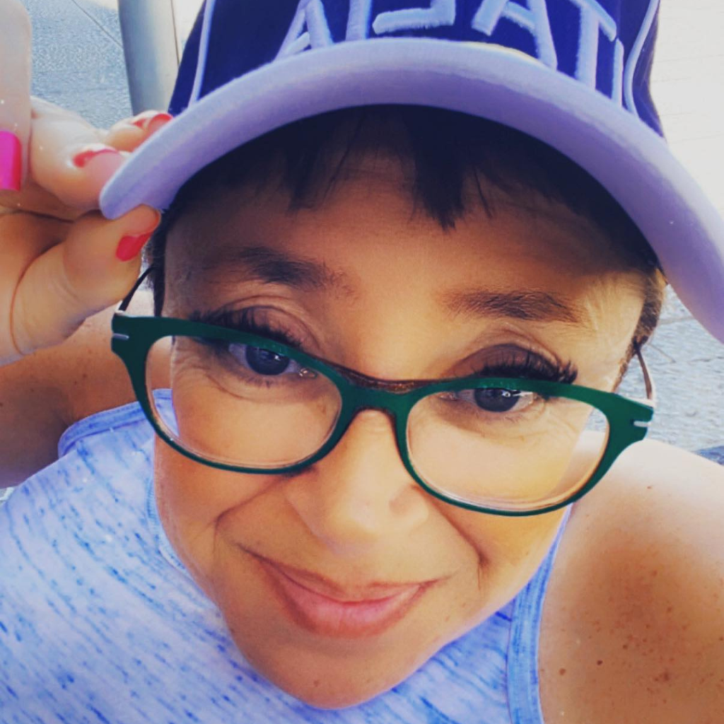 upclose photo of Lisa, a brunette female with green glasses on. She's wearing a baseball cap. She's holding it with her right hand. She's smiling. 