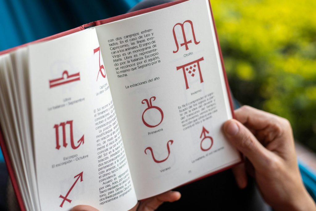 a book held open by white hands. the book has horoscope signs and text that isn't readable.