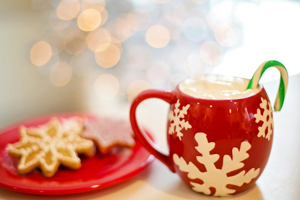 red plate with festive snowflake shaped cookies next to a red mug with snowflakes design and a candy cane in the hot cocoa.