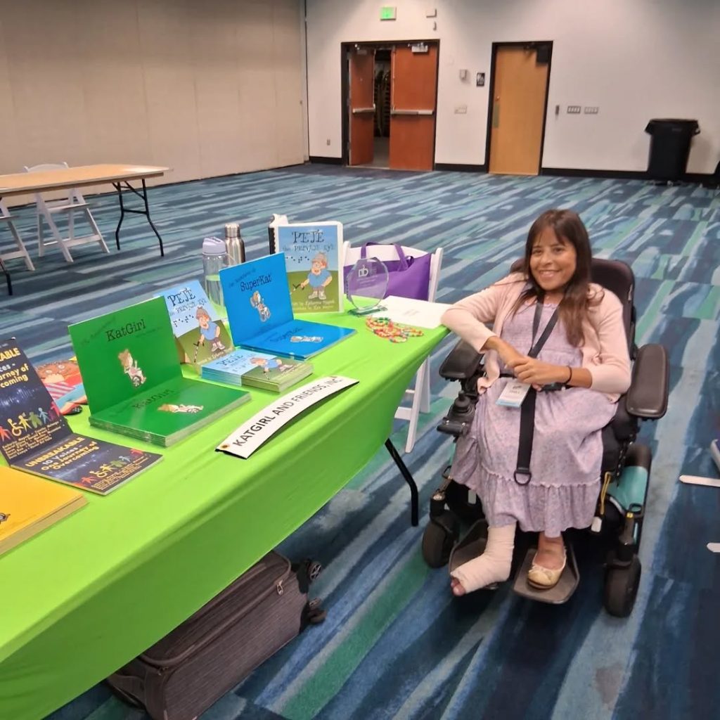 audacitymagazine.com guest writer, Katherine Magnoli is on the right side in her wheelchair. She is wearing a lavender dress. On the left is a long conference table with lime green colored table cloth showcasing her books.