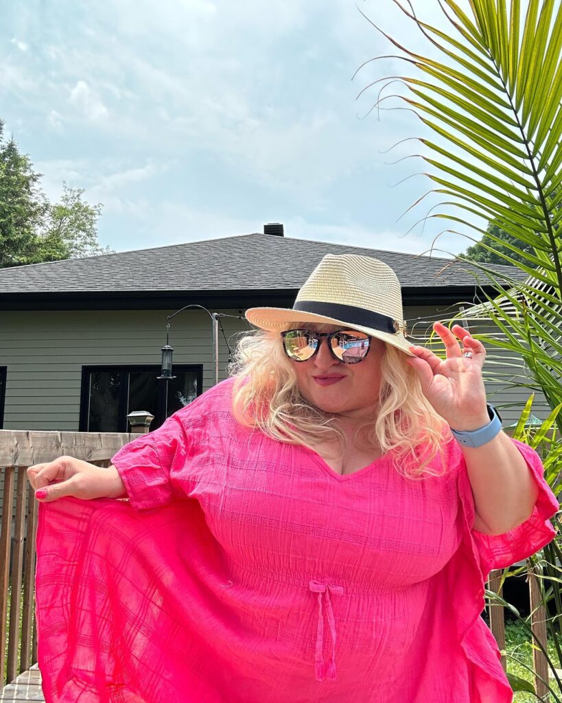 blonde female with sunglasses and straw hat. She's wearing a beautiful pink fuchsia dress. She's smiling at the camera reflected in the photo.
