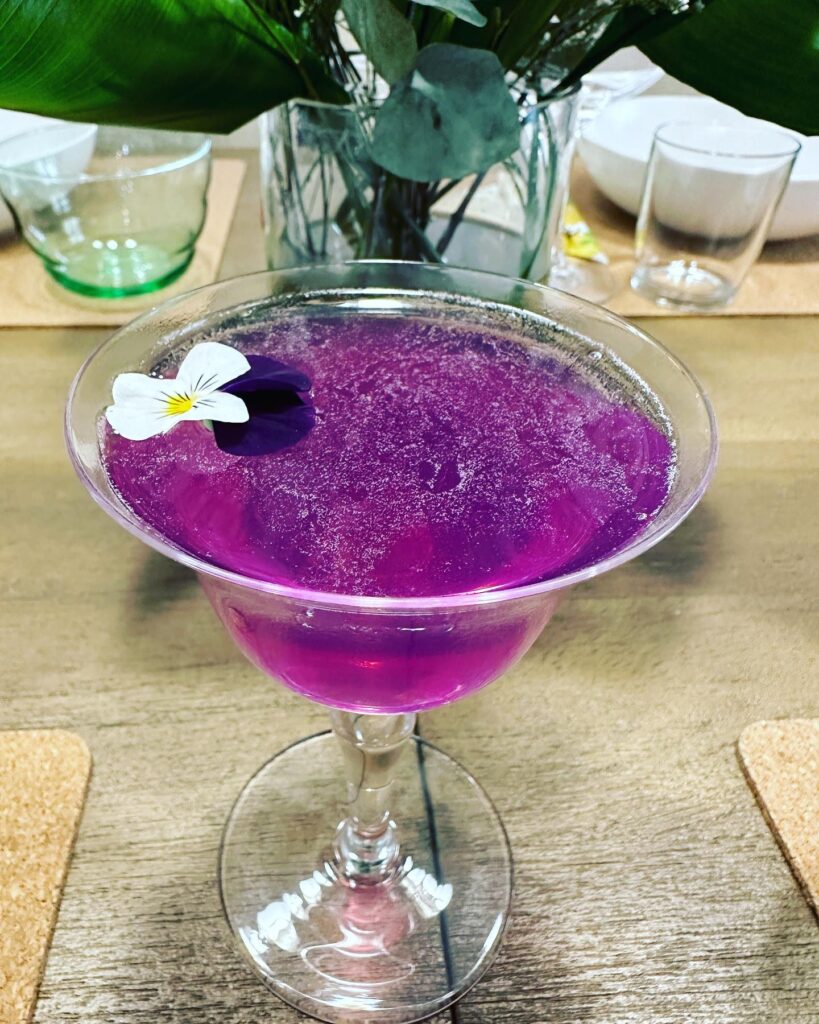 violet color cocktail with a white and purple edible flower. 