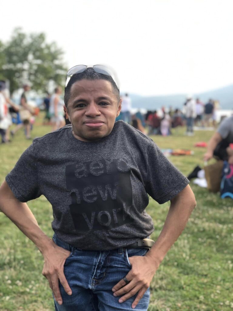 Rich, Latino man posing standing up for the camera. He's at the park. He's wearing jeans, gray t shirt, and has sunglasses on the top of his head.
