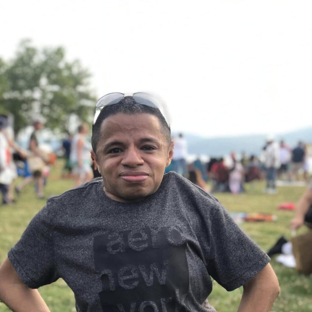 Rich, Latino man posing standing up for the camera. He's at the park. He's wearing jeans, gray t shirt, and has sunglasses on the top of his head.