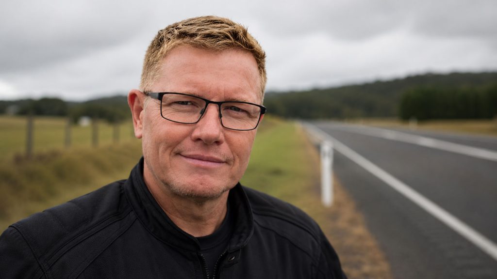 a selfie by a male with short blonde hair, square frame eye glasses. The background is blurry but it's a road with grass behind him.