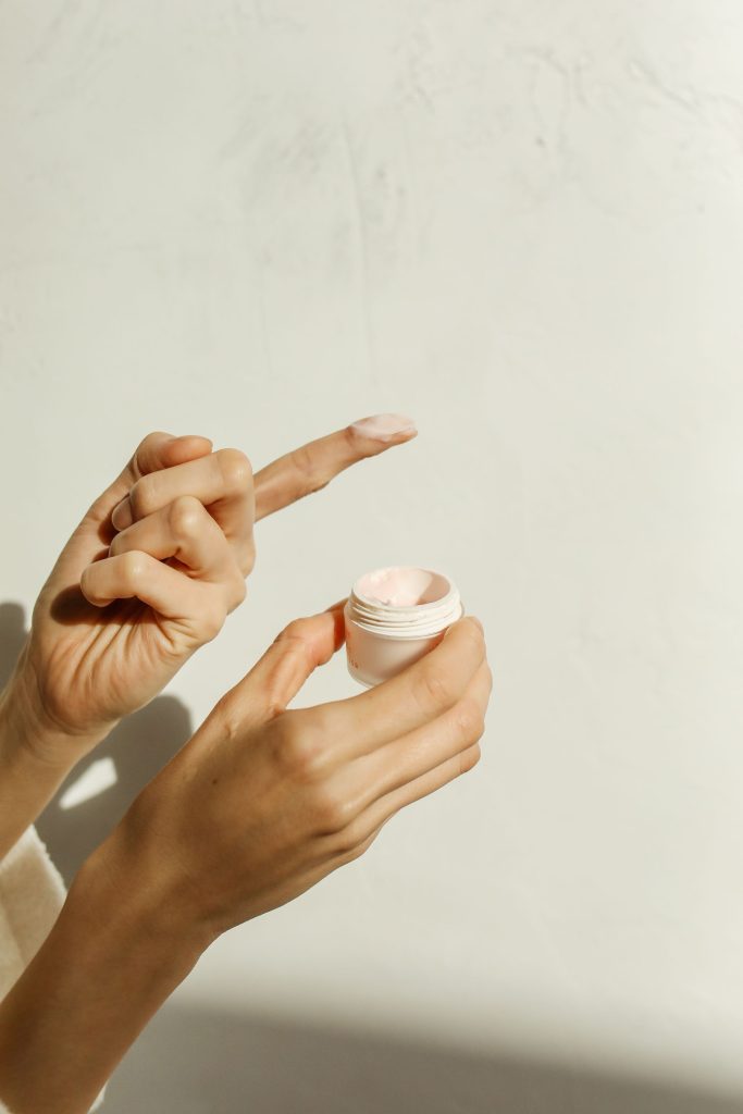 skin care image of a white female's hands. the right hand holds a small cream container and the left hand has the pointer finger extended with a dab of cream on it.