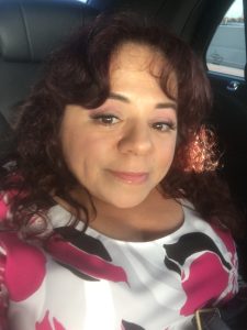 Latina author, Nathasha Alvarez smiles in her selfie. Dark brown hair with red tones shining from the sun. She's wearing a white, black, and pink top.