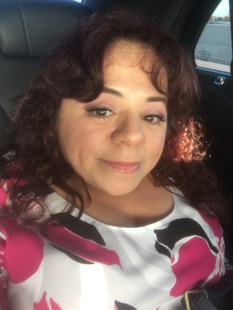 audacitymagazine.com
Latina author, Nathasha Alvarez smiles in her selfie. Dark brown hair with red tones shining from the sun. She's wearing a white, black, and pink top.